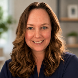 Smiling woman with wavy brown hair in a navy blouse, indoor background blurred into soft greens and grays.