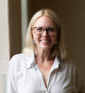 Smiling woman with blonde hair wearing tortoiseshell glasses and a light striped shirt, standing by a light wall.” ,
