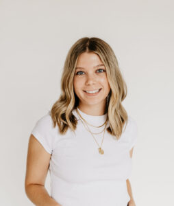 Smiling woman with blonde hair wearing a white t-shirt and blue jeans, standing against a light neutral background.