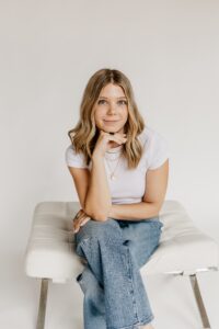 Young woman with blond hair sitting on a white chair, resting chin on her hand and smiling slightly against a neutral background, wearing a white t-shirt and blue jeans.