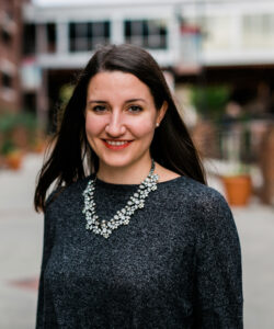 Smiling woman with dark hair wearing a sparkly statement necklace, outdoors in an urban plaza with blurred buildings behind her.