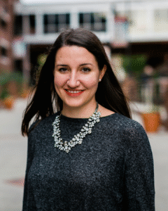 Smiling woman with dark hair in a gray knit sweater and a jeweled necklace, outdoors in an urban setting.