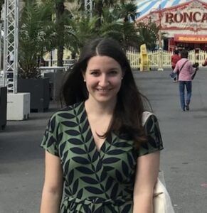 Smiling woman with long dark hair in a green leaf-patterned dress, carrying a light tote, at an outdoor fair with palm plants in the background.